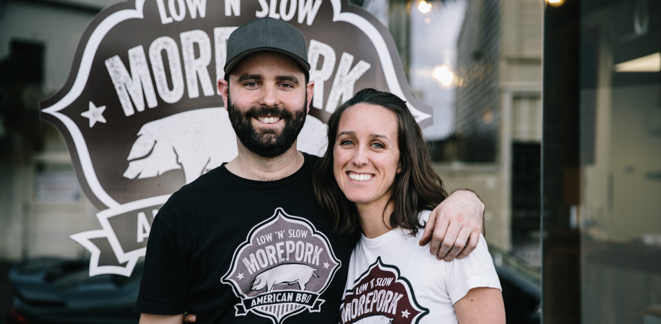 Two people standing in front of a 'Low N' Morepork' sign.